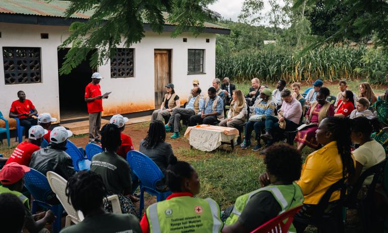 A group of people sits outdoors in front of a building, listening to a speaker in a red shirt, with some participants wearing uniforms and others taking notes.