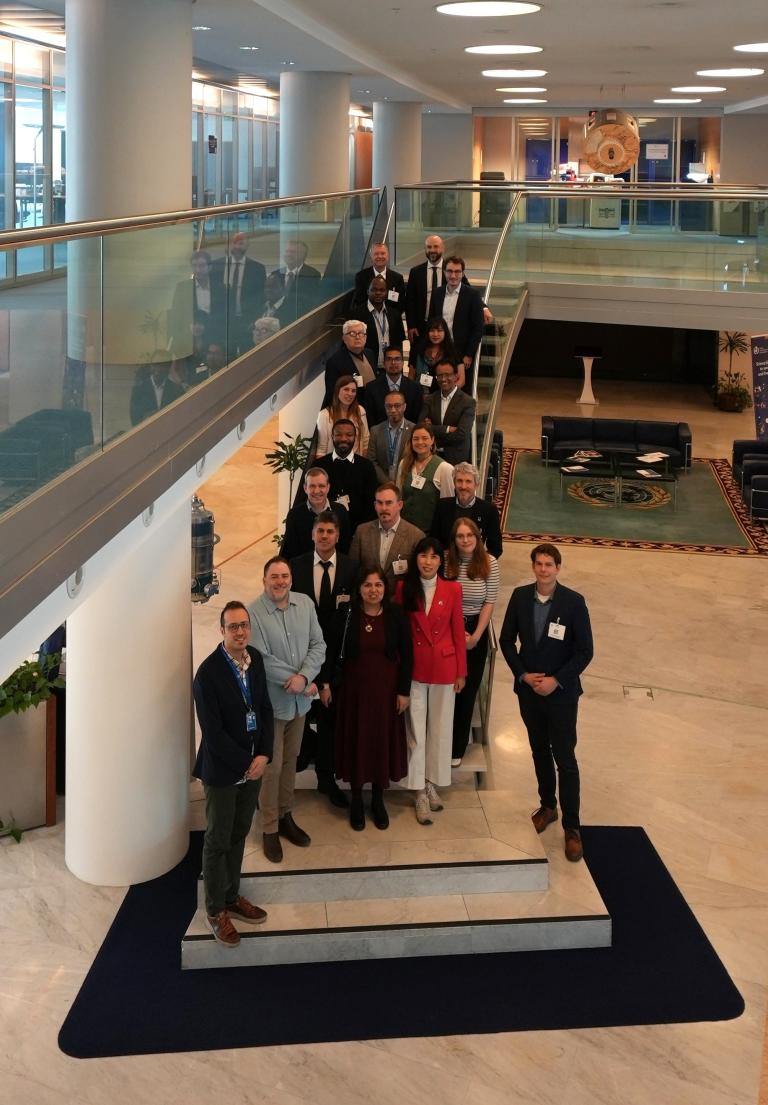 A group of people pose for a photo on a staircase in a modern building lobby with large windows and contemporary furnishings.