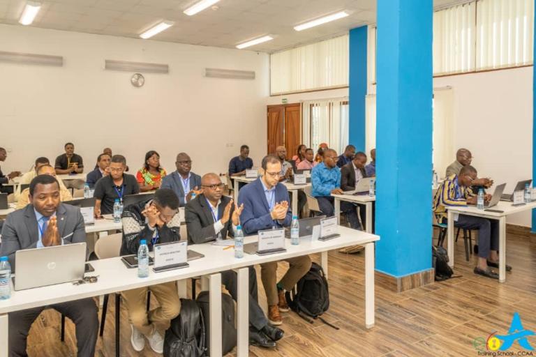 A group of people sit at desks with laptops and nameplates in a conference room, listening attentively. Some are clapping. Bottled water is on the tables.