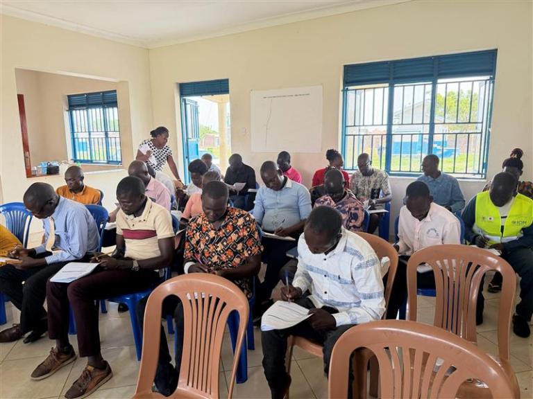 A group of adults sits in a classroom on plastic chairs, writing on papers, with windows and a whiteboard visible in the background.