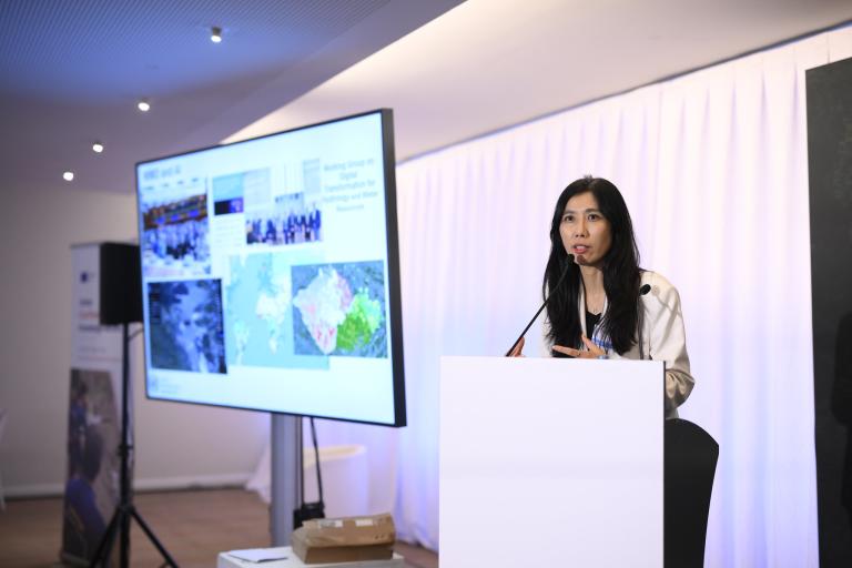A woman stands at a podium giving a presentation, with a large screen behind her displaying maps, images, and text.