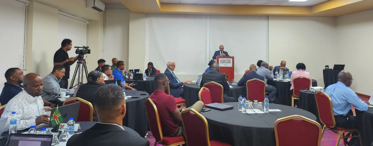 A speaker addresses a group of people seated at round tables in a conference room. A cameraman records the event while attendees listen and take notes.