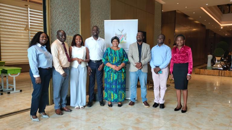 Eight people stand in a row indoors, posing for a group photo in front of a banner. They are dressed in business casual and formal attire.