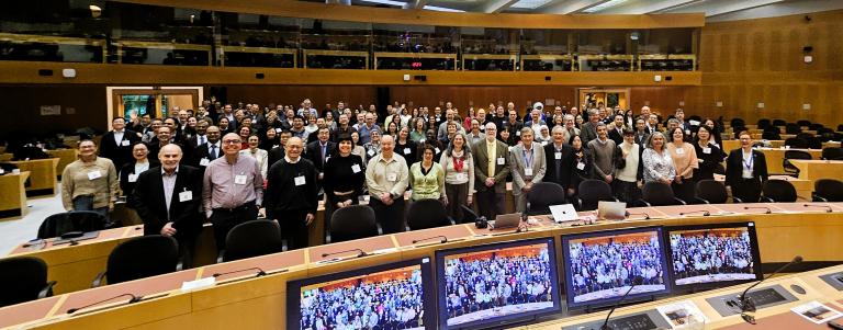A large group of people pose for a photo in a conference room, with monitors in the foreground displaying the same scene.
