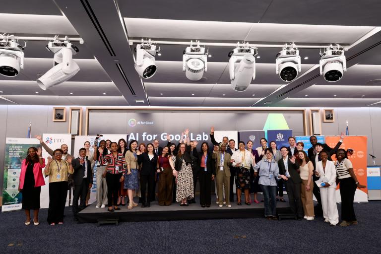 A large group of people stand together on a stage, posing for a photo at an event with "AI for Good Lab" displayed on a screen behind them.