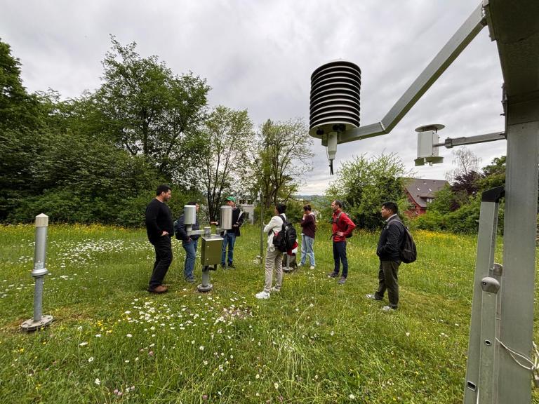A group of people stands in a grassy field examining weather instruments, including a Stevenson screen and rain gauges, surrounded by trees under a cloudy sky.
