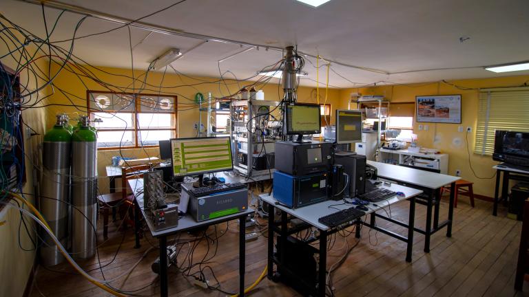 Laboratory room with scientific equipment, computers, gas cylinders, and numerous wires connected to devices on tables, with yellow walls and wooden floor.