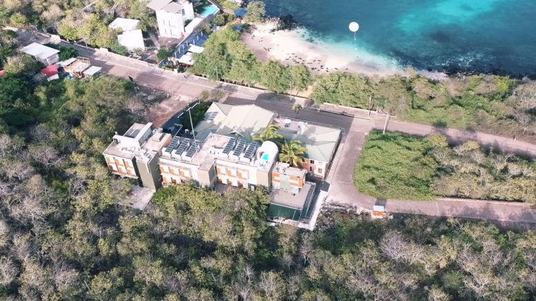 Aerial view of a modern building surrounded by trees, with a road, parked cars, and a beach with turquoise water in the background.