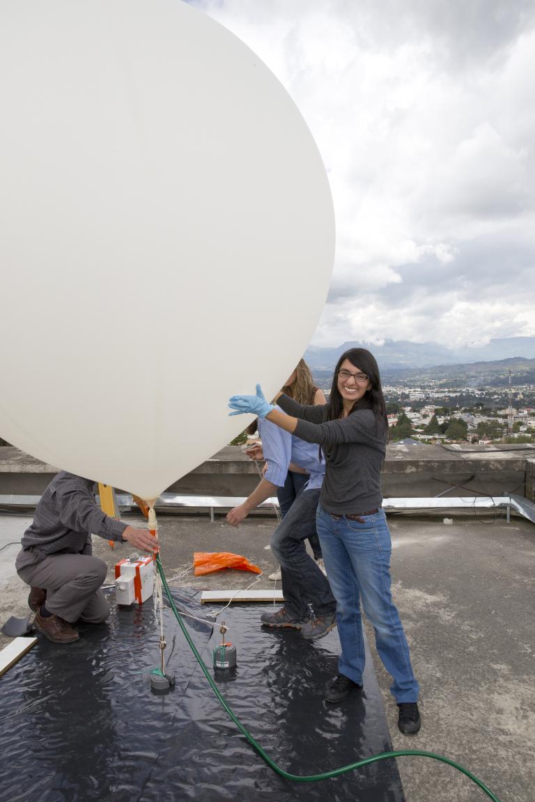 A woman in gloves smiles at the camera while holding a large white weather balloon on a rooftop, with two other people working beside her.
