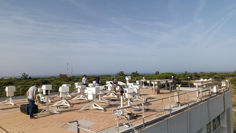 Several people work among multiple white scientific instruments mounted on tripods on a rooftop, with trees and the sea visible in the background under a clear sky.