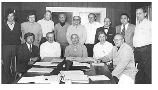 A group of fourteen men, some seated at a table with papers, others standing behind them, pose for a black-and-white photo in an office setting.