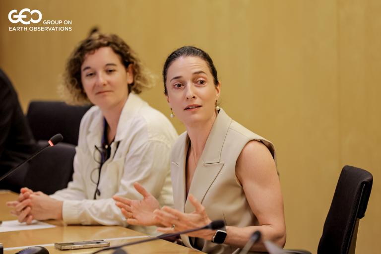 Two women sit at a conference table, one speaking while the other listens. Both have papers and a microphone in front of them. The background features a plain wooden wall.