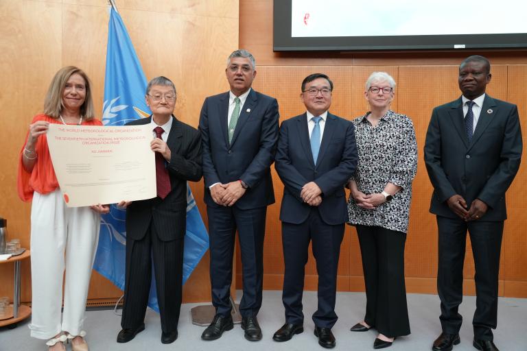 Six people stand indoors; one person on the left holds a large certificate. A United Nations flag is visible in the background.