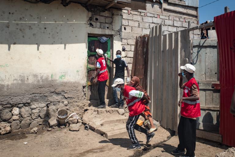 Red Cross workers, some wearing masks, assist people outside a modest building with stone and brick walls in a rural setting.