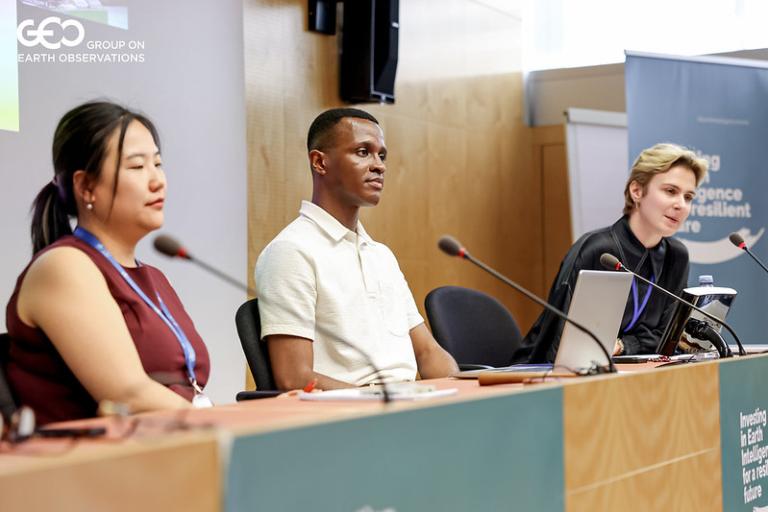 Three panelists sit at a conference table with microphones, participating in a discussion at a professional event.