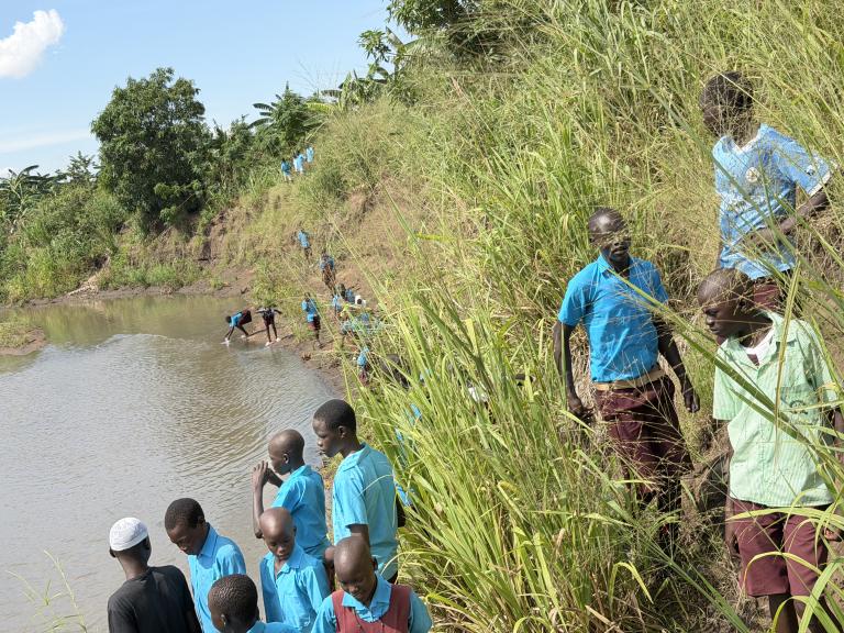 A group of students in blue uniforms walk along a grassy riverbank, with some standing near the water and others climbing the slope.