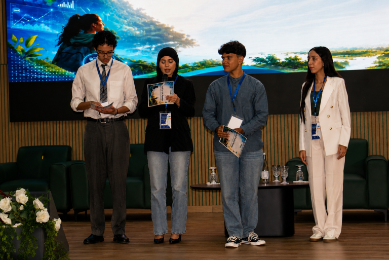 Four people with conference lanyards stand on a stage holding booklets, with a large nature-themed screen behind them and chairs in the background.
