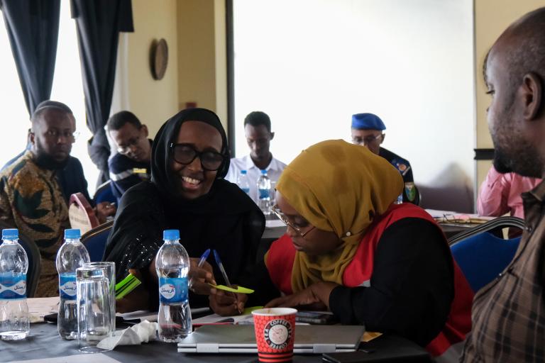 Several people sit around a table with water bottles, two women in the foreground discussing documents, one smiling and holding a pen. Other attendees are visible in the background.
