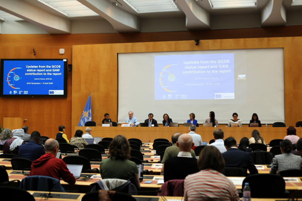 Panelists sit at a long table addressing an audience in a conference room, with a blue presentation slide displayed on large screens at the front of the room.