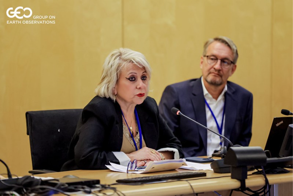 Two people sit at a conference table with microphones and laptops; one woman is speaking while the man listens. A “GEO Group on Earth Observations” logo appears in the top left corner.