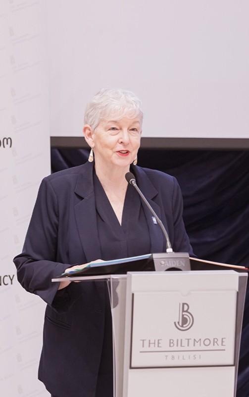 An older woman with short gray hair speaks at a podium labeled “The Biltmore Tbilisi,” wearing a dark blazer and earrings, in front of a backdrop with text.