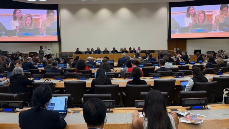 A large conference room with attendees seated facing a panel of speakers at the front, two large screens display the panelists.
