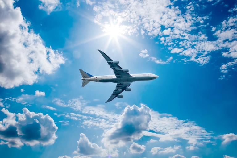 A large airplane flies in a bright blue sky with scattered clouds and sunshine above.