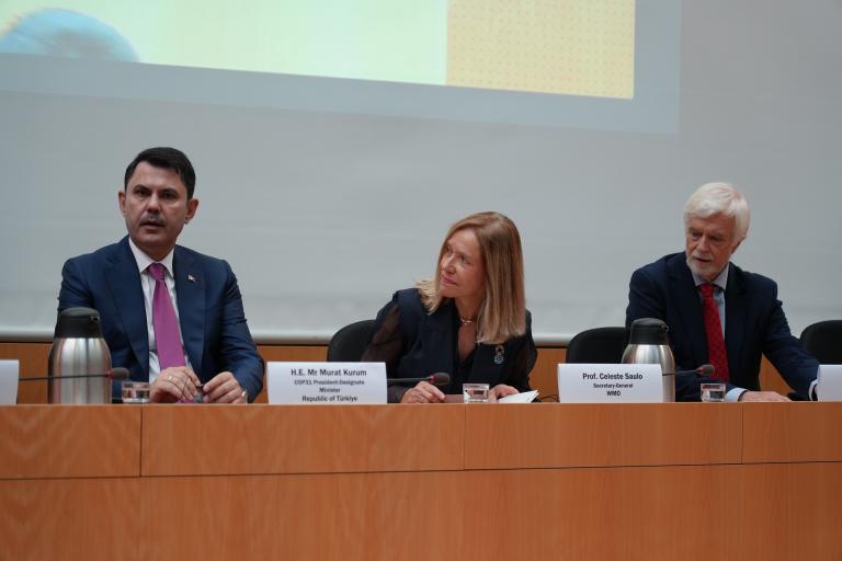 Three people in formal attire sit at a panel table with microphones and nameplates during a conference or meeting.