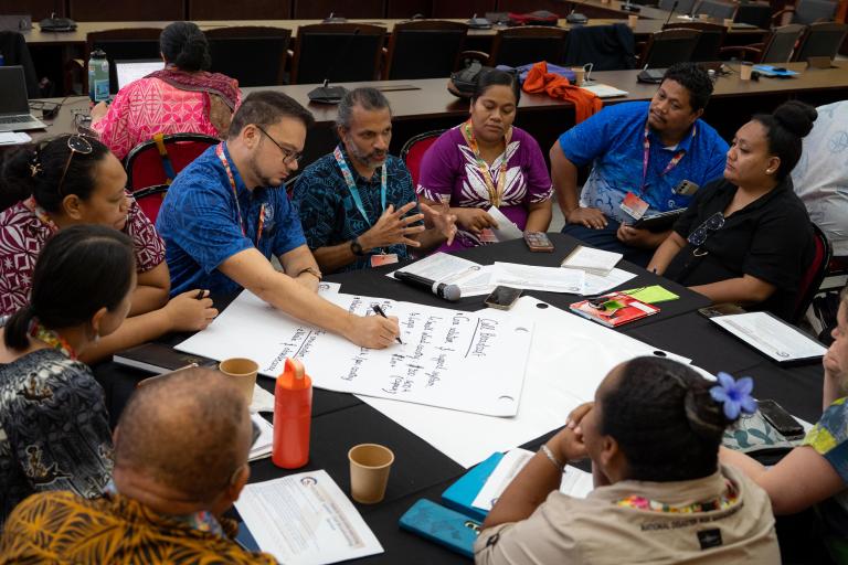 A group of people sit around a table in discussion, with one person writing on a large sheet of paper and documents, cups, and a phone on the table.