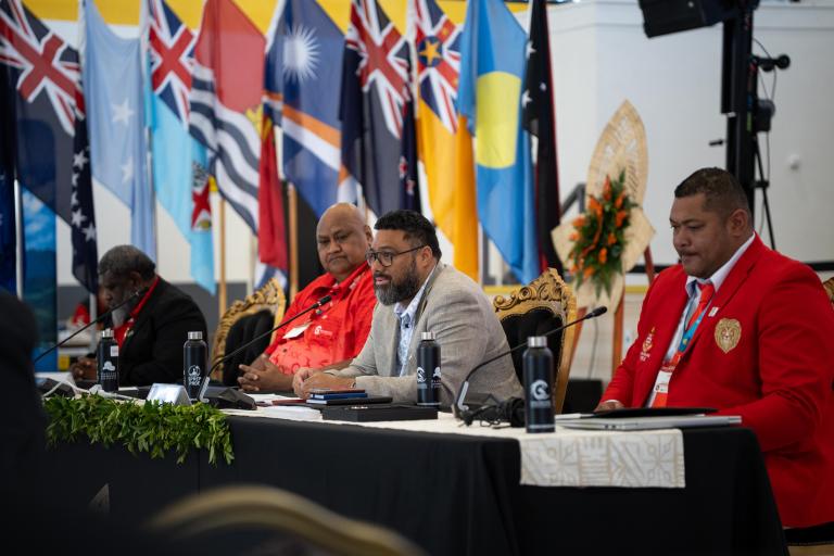 Four men sit at a conference table with microphones and documents; various national flags are displayed in the background.