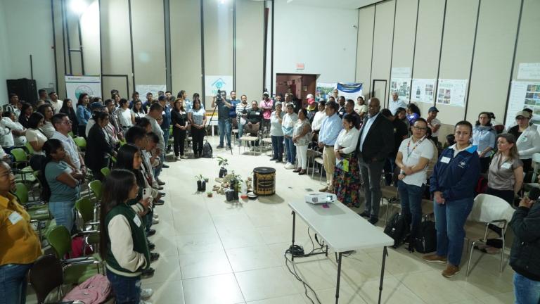 A large group of people stands in a conference room, facing each other in a circle around a display of plants and objects at the center. Posters are visible on the walls.