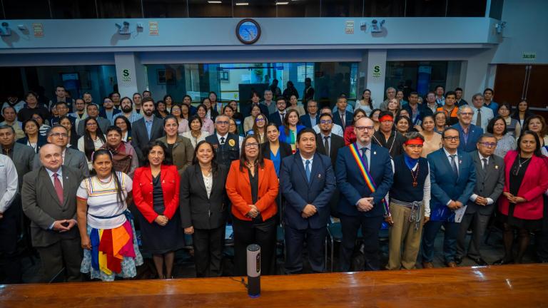 A large group of people, including men and women in formal and traditional attire, stand together in a conference room, facing the camera.