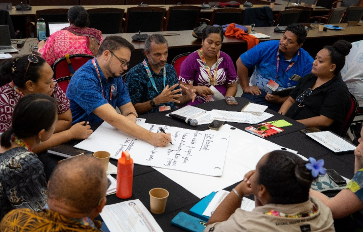 A group of people sit around a table in discussion, with one man writing on a large sheet of paper and others listening and taking notes.