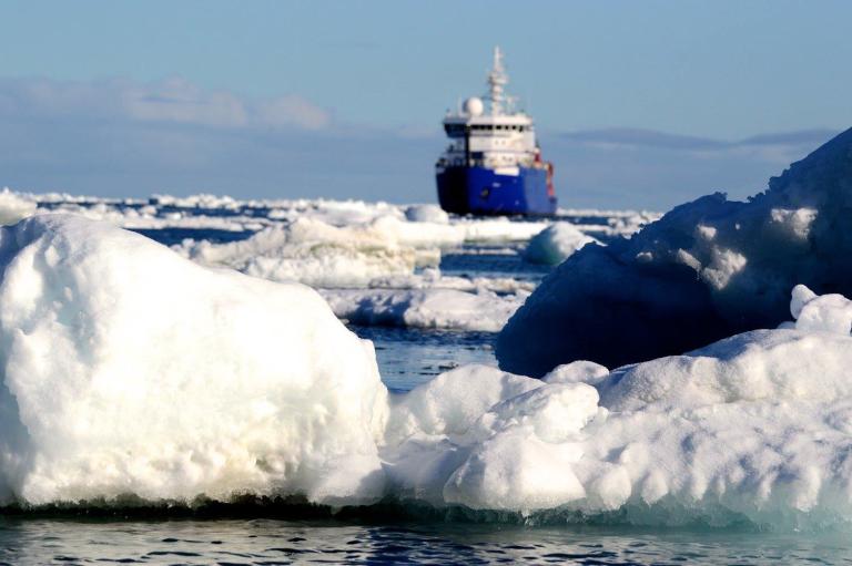 A large ship sails through icy waters, surrounded by floating icebergs and snow under a clear blue sky.