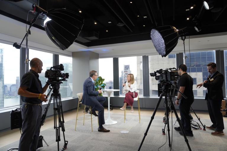 A man in a suit and a woman in a white blazer are interviewed in a high-rise office with camera crews and lighting equipment.