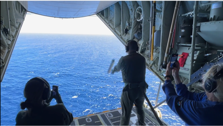 Crew members in a cargo plane doorway looking out over the ocean as one person tosses an object into the water.