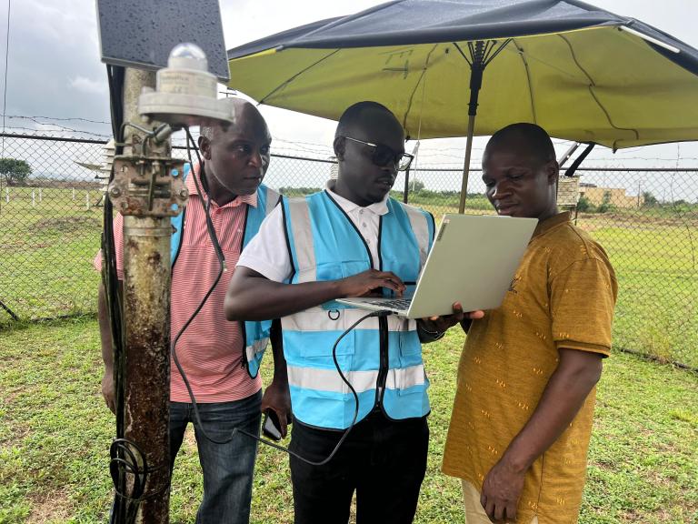 Three men stand outdoors beside meteorological equipment; one in a blue vest uses a laptop under a large umbrella, while the other two observe. A chain-link fence is in the background.