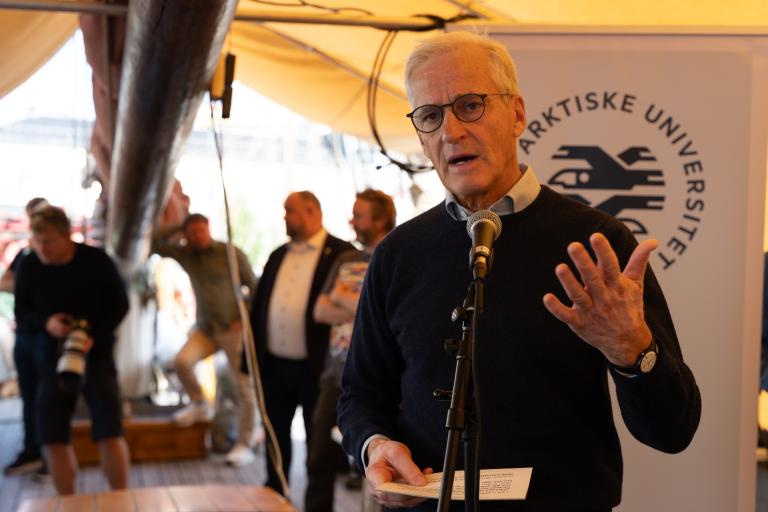 A man with grey hair and glasses speaks into a microphone while holding a paper, with the UiT The Arctic University of Norway logo visible on a sign behind him.