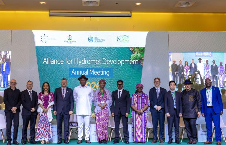 Thirteen people stand in a row on stage in front of a projection screen for the Alliance for Hydromet Development Annual Meeting.
