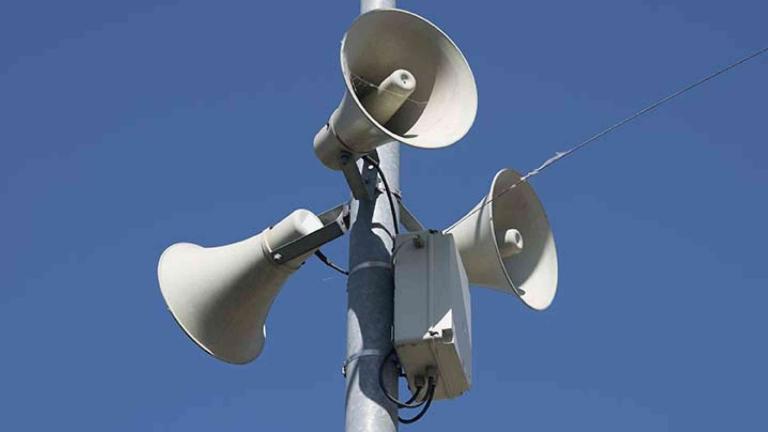 Three loudspeakers mounted on a metal pole against a clear blue sky.