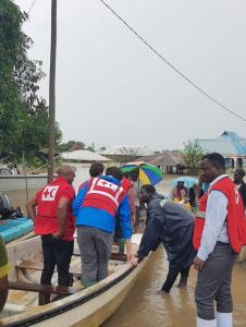 People in life vests assist others into a boat in a flooded urban area with trees and buildings visible in the background.