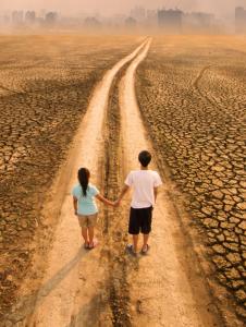 Two people stand on a dry, cracked path leading toward a distant city skyline under a hazy sky.
