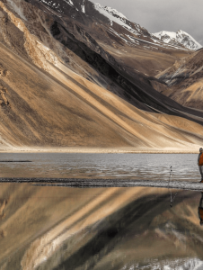 A person stands near a tripod by a reflective mountain lake with snow-covered peaks in the background.