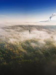 Aerial view of a forest covered in mist, with sunlight shining and smoke rising from an industrial facility in the background.