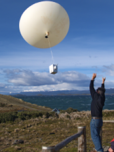 Three people outdoors launch a large white weather balloon with a box attached, near a wooden fence and open landscape under a blue sky.