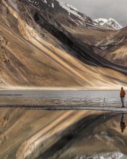 A person stands near a tripod by a reflective mountain lake with snow-covered peaks in the background.