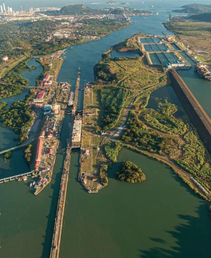 Aerial view of the panama canal with multiple lock gates open, connecting large bodies of water, surrounded by lush greenery and infrastructure.