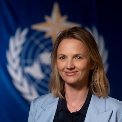 A person with shoulder-length blonde hair wearing a light blue blazer smiles in front of a blurred background featuring a blue flag with a white emblem.