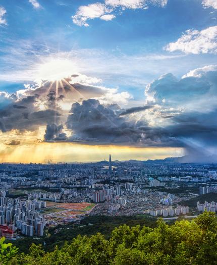 Panoramic view of a cityscape with dramatic clouds and sun rays breaking through. A rainstorm is visible in the distance, surrounded by green hills and urban buildings under a blue sky.