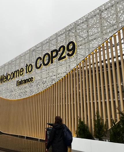 Entrance design for COP29 with a sign that reads "Welcome to COP29 Entrance," featuring wooden slats and a decorative perforated metal surface. People are walking nearby.